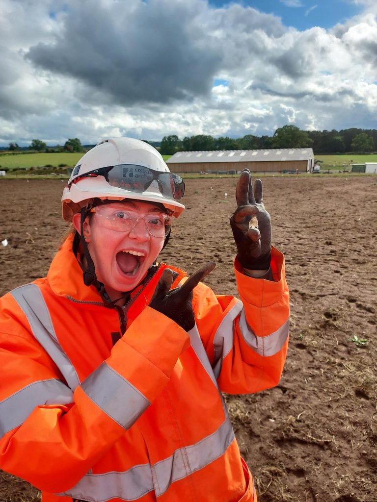 Archaeologist in orange hi-vis jacket standing in a field holding a stone arrow head.